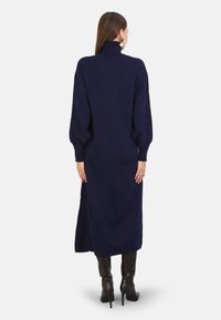 Abito lavorato a maglia blu navy con colletto alto, maniche a palloncino lunghe e silhouette dritta, che arriva fino al polpaccio. Indossato con stivali alti fino al ginocchio.
