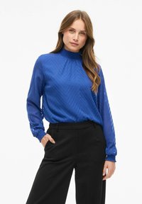 Donna con capelli lunghi, vestita con una blusa blu a maniche lunghe dalla trama e pantaloni neri a vita alta, in piedi con una mano in tasca.