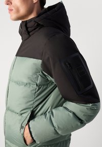 Pufferjack met een zwarte капюшон en bovenkant, groene onderkant, zijzakken en een ritszak op de mouw met logo-details.