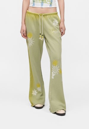 Person iført lysegrønne vidde sweatpants med solmotiver og snøre i taljen, kombineret med hvide sko og en cropped top.