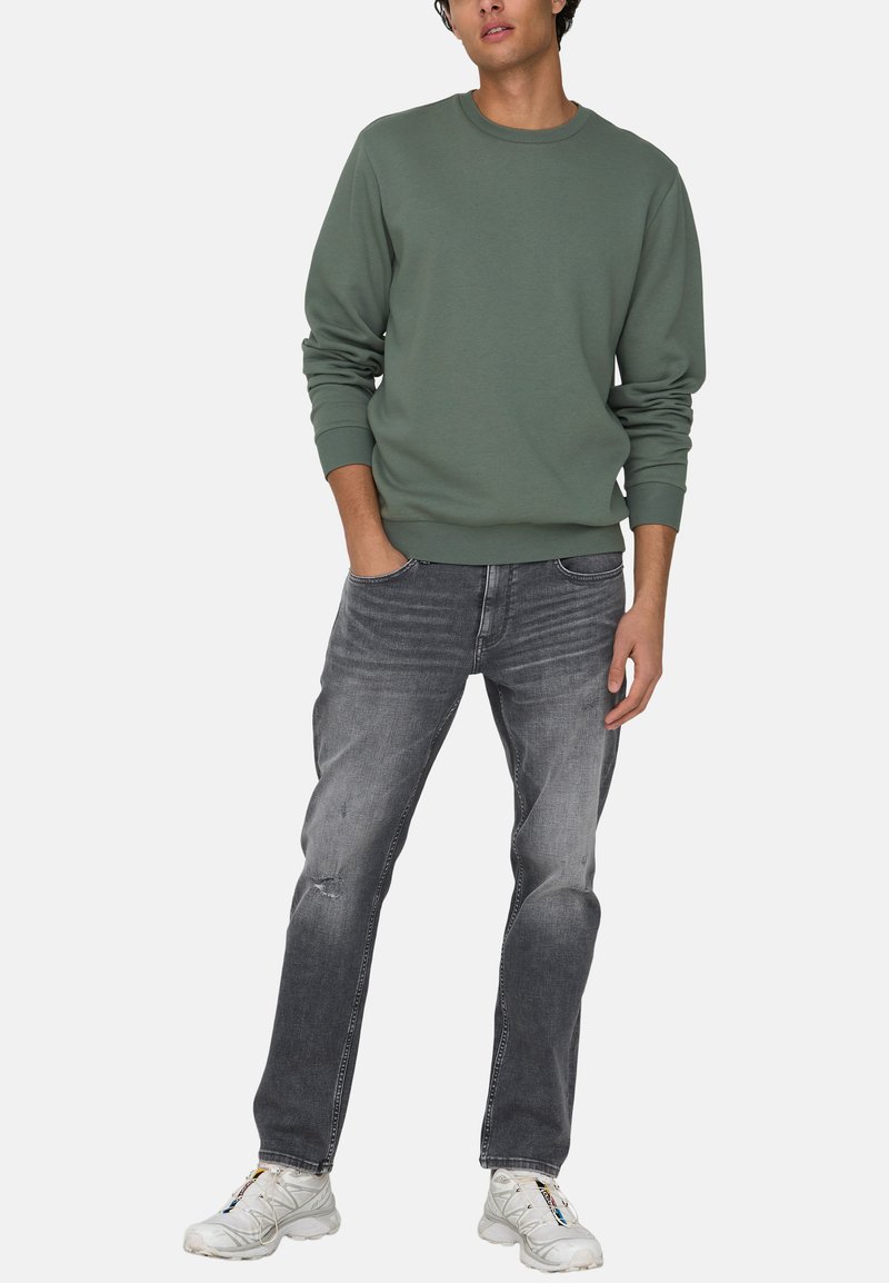 Vihreä puuvilla-sweatshirt pitkillä hihoilla, yhdistettyinä haalistuneisiin harmaisiin farkkuihin ja valkoisiin lenkkareihin. Asu on rento ja yksinkertainen muotoilu.