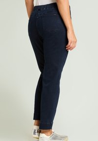 Donna che indossa pantaloni slim-fit blu scuro e sneakers bianche, in piedi con una leggera vista laterale e posteriore su uno sfondo chiaro.