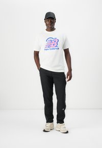 Hvid kortærmet t-shirt med et blåt og lyserødt grafisk design, sorte slim-fit bukser og beige sneakers med grå detaljer. Sort kasket.