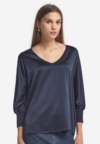 Navyblaue Satinbluse mit V-Ausschnitt, langen Puffärmeln und glatter Textur. Der Saum ist hinten leicht länger.