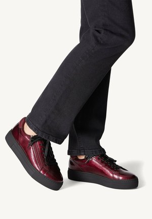 Person wearing black pants and shiny dark red sneakers with black laces and thick black soles, standing on a light background.