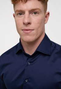 Granatowa koszula zapinana na guziki wykonana z gładkiego materiału. Posiada wskazujący kołnierzyk oraz dwa widoczne guziki na przedniej listwie.