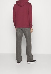 Personne portant un sweat à capuche bordeaux, un jean gris ample et des baskets noir et blanc, debout dos au spectateur sur un fond uni.