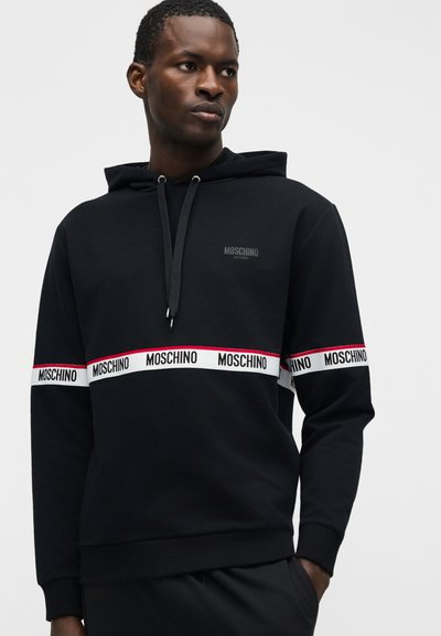 Άνδρας που φοράει μαύρο φούτερ Moschino με λευκή και κόκκινη ρίγα και επαναλαμβανόμενο λογότυπο "Moschino" γύρω από το στήθος και τα μανίκια, με χέρια στις τσέπες.