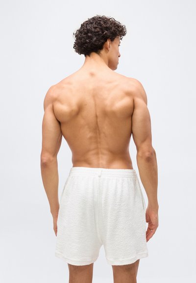 Homme musclé aux cheveux bouclés, debout de dos, portant un short blanc texturé sur un fond clair uni.