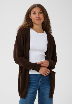 Femme aux cheveux bouclés portant un cardigan marron foncé, une chemise blanche et un jean bleu, debout devant un fond blanc uni.