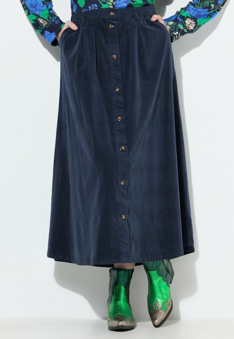 Marineblauwe, knielange rok van katoen met plooien en voor knopen. Gecombineerd met groene metallic enkellaarzen met decoratieve accenten.