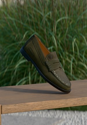 Donkergroene suède herensloffer met stikseldetails, pennyriem en reliëflogo, getoond op een houten oppervlak met groene planten op de achtergrond.