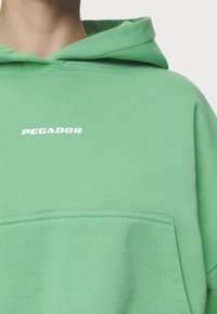 Sudadera verde con un bolsillo frontal, hecha de tela suave. Presenta un logo blanco "PEGADOR" en el pecho y una capucha con cordones.