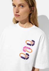 Biała bawełniana koszulka z kolorowymi haftowanymi akcentami w kolorze różowym, fioletowym, pomarańczowym i beżowym. Luźny krój z okrągłym dekoltem.