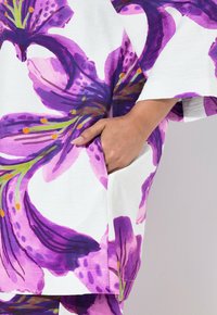 Blomstretrykt top med store lilla og pink liljemønstre på en hvid baggrund, med en sidelomme og afslappet, løst fit.