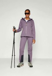 Purple hiking jacket with zip, paired with matching pants featuring black accents. Model holds trekking poles, wearing dark sunglasses and hiking shoes.