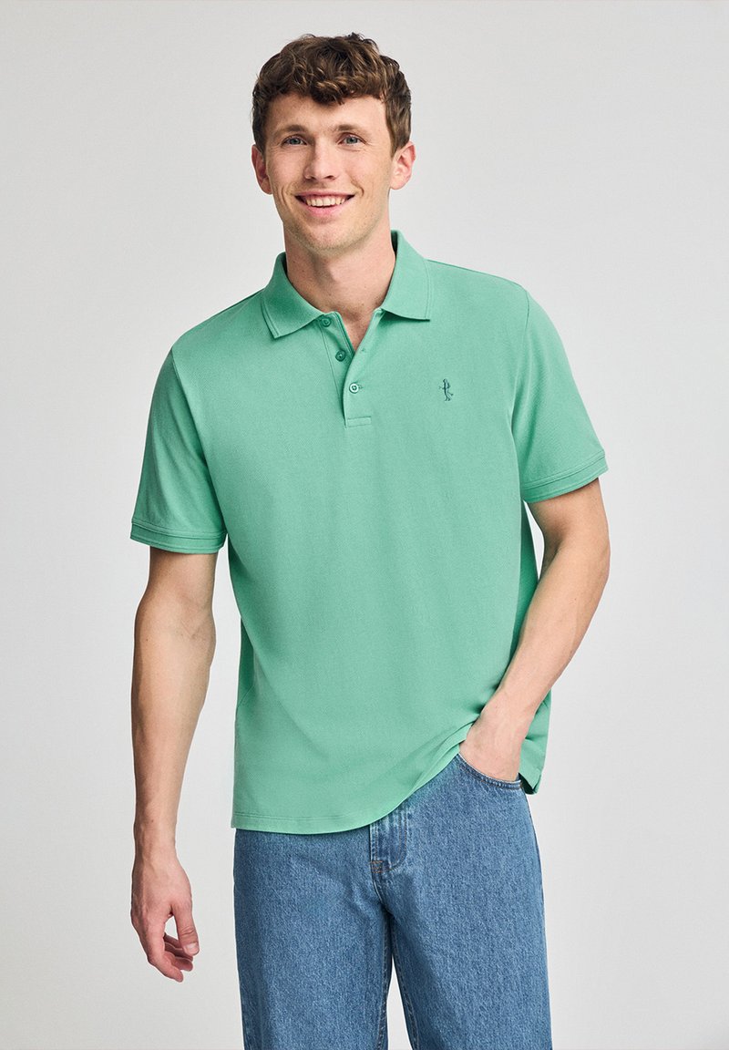Camisa polo verde menta de manga corta, con cuello clásico y logo bordado. Combinada con jeans azul claros, con textura de tela suave.