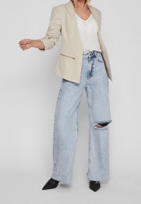 Blazer beige con bolsillo con cremallera, camiseta blanca de cuello en v, vaqueros de pierna ancha azul claro con rodilla rasgada y tacones negros puntiagudos.