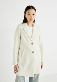 Blazer bianco su misura con rever a notch, due bottoni e tasche laterali; realizzato in un tessuto testurizzato, abbinato a un top semplice e denim.