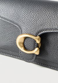 Μαύρο πορτοφόλι από δερμάτινη υφή με κούμπωμα σε σχήμα πετάλου από μέταλλο σε χρυσό τόνο σε άσπρο φόντο.