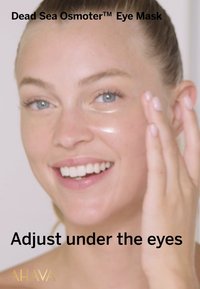 Transparent gelögonmask, applicerad under ögonen, med slät textur. Användarens hand placerad på masken, ljus hudton och ett klart leende.