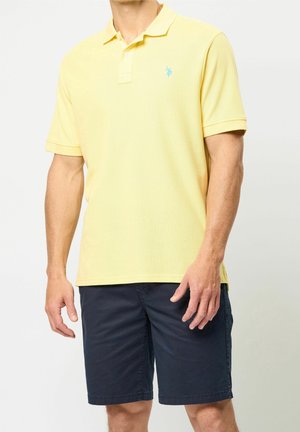 Mann trägt ein hellgelbes kurzärmeliges Poloshirt und dunkelblaue knielange Shorts vor einem schlichten Hintergrund.