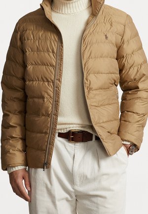 Mężczyzna w beżowej pikowanej kurtce na kremowym golfie, białe spodnie sztruksowe oraz brązowy skórzany pasek i zegarek.