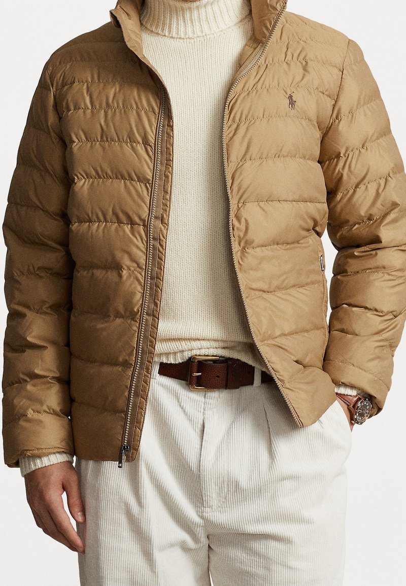 Mand iført tan quiltet jakke over cremefarvet rullekravetrøje med hvide kordfløjlsbukser og brunt læderbælte samt ur.