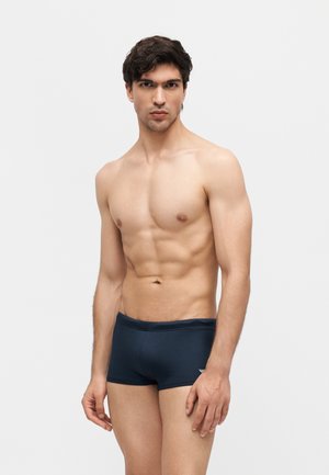 Izmos férfi rövid sötét hajjal, sötétkék fürdőnadrágot visel, egy egyszerű világosszürke háttér előtt áll.