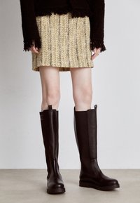 Brązowe kozaki sięgające kolan, wykonane z gładkiej skóry, z gumową podeszwą i bocznymi ozdobami. Noszone z teksturowaną spódnicą w żółto-czarną kratę tweedową.