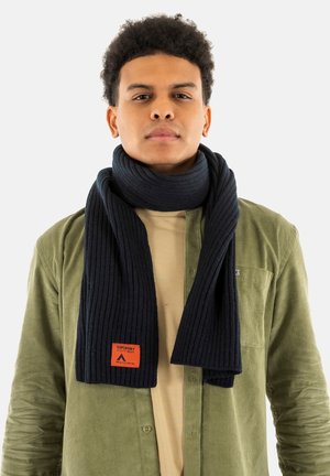 Scarf a costine di un blu scuro, realizzata in un tessuto morbido, con una toppa quadrata arancione che presenta il logo del brand. Indossata sopra una maglietta beige chiaro e una giacca verde oliva.