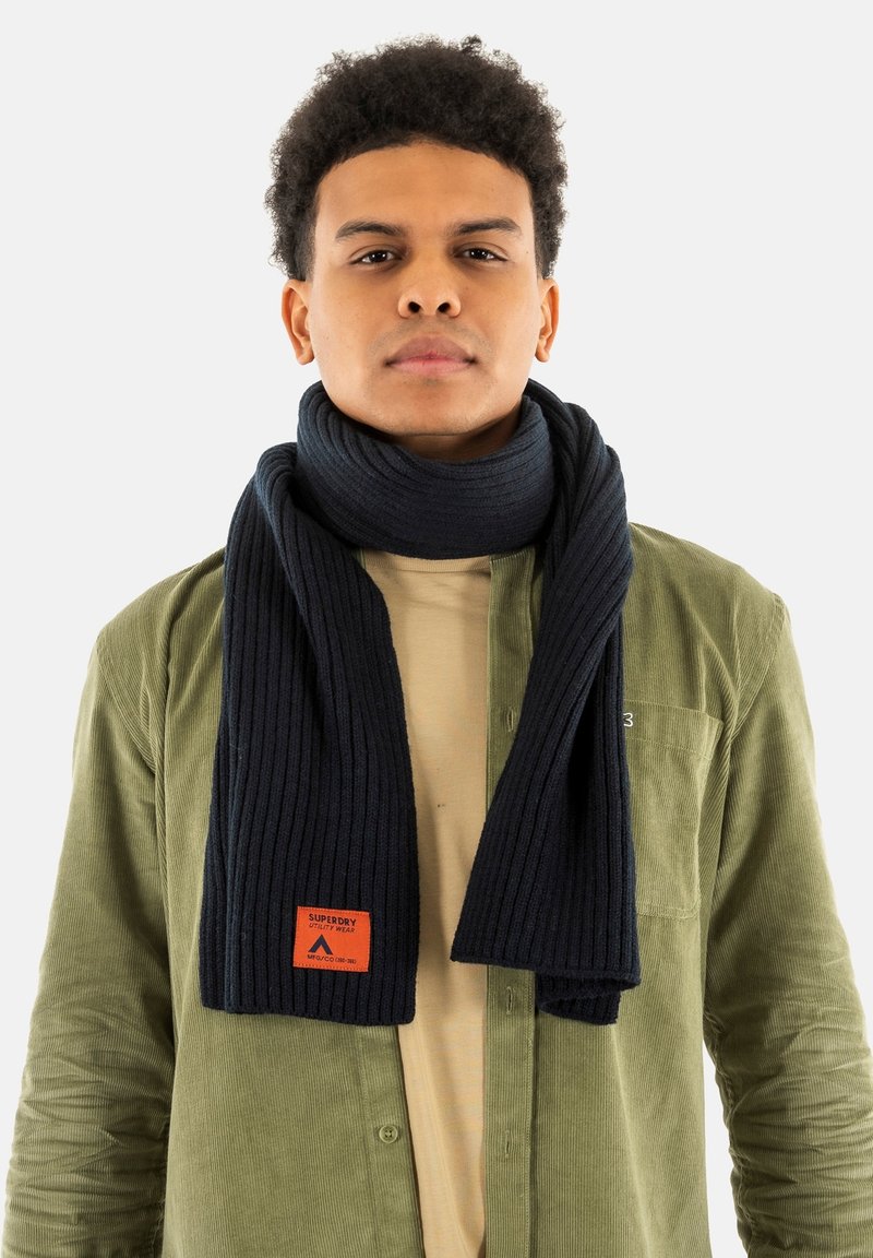 Écharpe côtelée bleu marine foncé en tissu doux, ornée d'un patch carré orange avec le logo de la marque. Portée sur un haut beige clair et une veste verte olive.