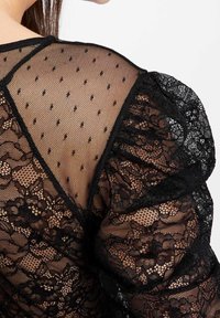 Schwarze Spitzenbluse mit einem Netzüberzug und kleinen Punkten, Rüschensäumen und floralen Spitzenakzenten auf dem Rücken.