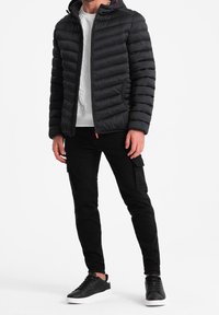 Sort pufferjakke med horisontalt quiltet mønster, lynlåslukning og hætte. Bæres over en hvid T-shirt, med sorte cargo bukser og sorte sneakers.