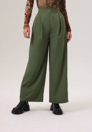 Personne portant un pantalon ample couleur olive, des bottes noires épaisses et un haut ajusté à manches longues avec un imprimé d'art de la Renaissance.