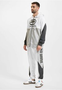 Sweat à capuche zippé gris et blanc avec logo, associé à un pantalon de survêtement gris et blanc arborant également un logo. Le modèle porte des sneakers blanches avec des rayures noires.