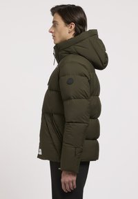 Chaqueta tipo plumón en verde oliva oscuro, con capucha, bolsillos con cremallera y un parche de logo. El material parece suave y aislante.