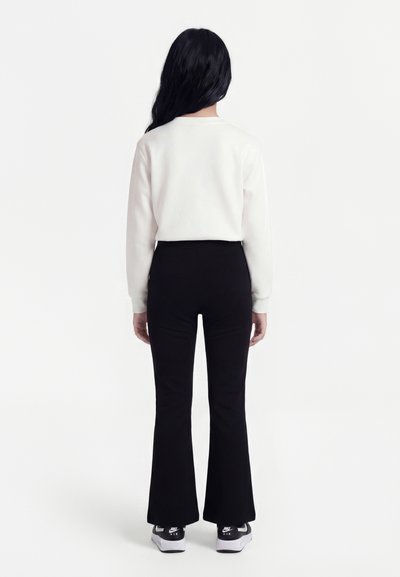 Personne debout de dos, portant un haut blanc à manches longues, un pantalon noir évasé et des baskets Nike Air noir et blanc.