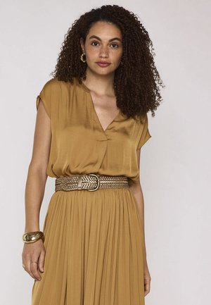 Femme aux cheveux bouclés porte un chemisier moutarde rentré dans une jupe plissée, ceinture tressée, boucles d'oreilles anneaux dorés et bracelet massif, debout devant un fond blanc.