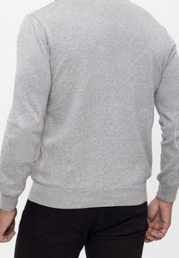 Homme portant un pull gris clair avec des coudières rembourrées et un pantalon foncé, vu de dos sur un fond uni.