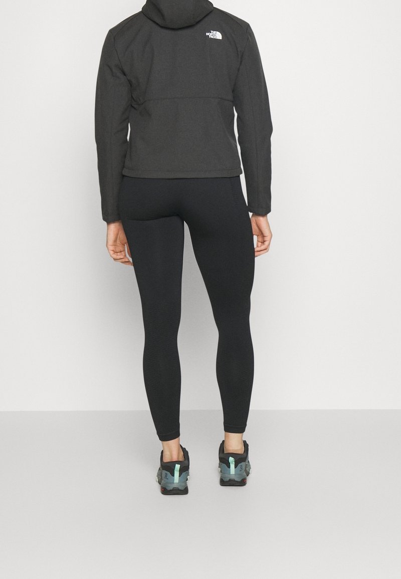 Giacca con cappuccio nera con logo sulla schiena, abbinata a leggings neri e scarpe da ginnastica grigie con dettagli turchesi, vista da dietro.