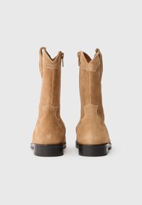 Stivaletti alla caviglia in suede marrone con una texture liscia, caratterizzati da un design curvato, chiusura con zip e dettagli di cuciture leggere sul retro.