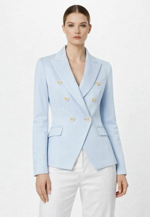 Donna che indossa un blazer doppiopetto su misura di colore azzurro chiaro con bottoni beige e pantaloni bianchi, in piedi davanti a uno sfondo semplice.