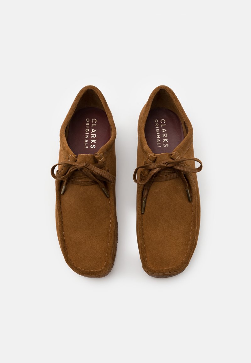 Brown suede shoes with a rounded toe, leather laces, and stitched detailing. The insoles feature "CLARKS ORIGINALS" branding in white.