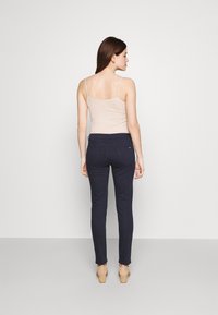 Marineblauwe fitted broeken met achterzakken, gecombineerd met een lichtbeige camisole. De stof heeft een gladde textuur; de schoenen zijn lichte hakken.