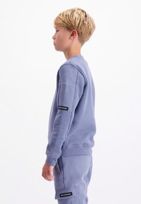 Jongen met blond haar in een bijpassende paarse sweatshirt en broek, staande in profiel tegen een witte achtergrond.