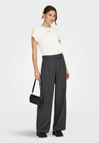 Femme portant un top blanc à manches courtes, un pantalon gris à jambes larges taille haute, des chaussures plateformes noires et tenant un petit sac à main noir.