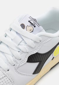 Hvit lærsneaker med svarte detaljer, med en teksturert sidepanel, lyseblå snøringer og et grafisk motiv av en karakter på tungen.