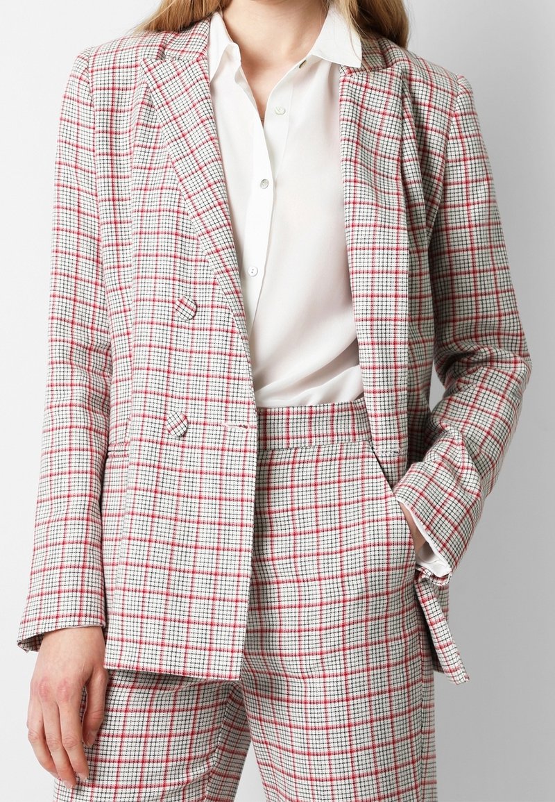 Kvinde iført en hvid skjorte under en rød og sort ternet blazer og matchende højtaljede bukser med den ene hånd i lommen.
