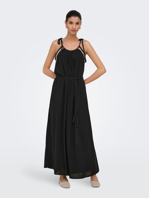Mujer de pie con un vestido maxi negro sin mangas con tirantes anudados en los hombros y un cinturón trenzado, con zapatos planos beige sobre un fondo liso.